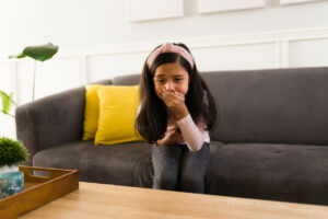 A sick and vomitting girl is sitting on the couch and looks like she is about to throw up.